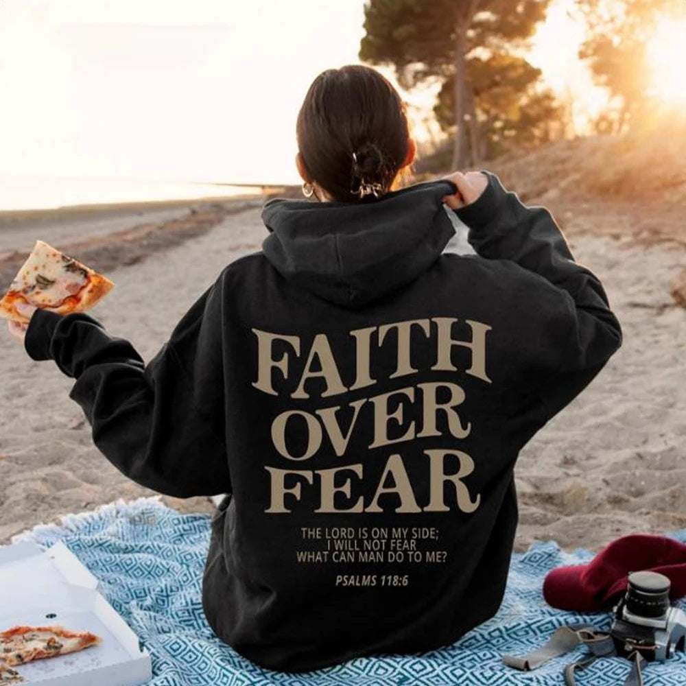 Woman wearing black Faith Over Fear Christian hoodie sitting on beach holding pizza slice at sunset