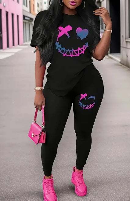 Woman wearing black T-shirt and leggings set with colorful smiley face design, paired with pink sneakers and matching handbag, standing on urban street.