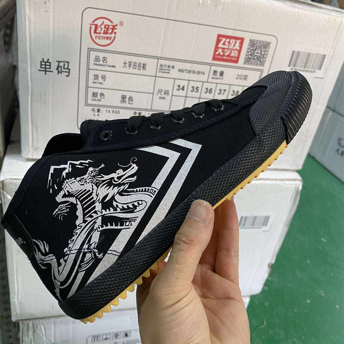 Black canvas mid-top sneaker with dragon design and gum sole held in hand against stacked boxes