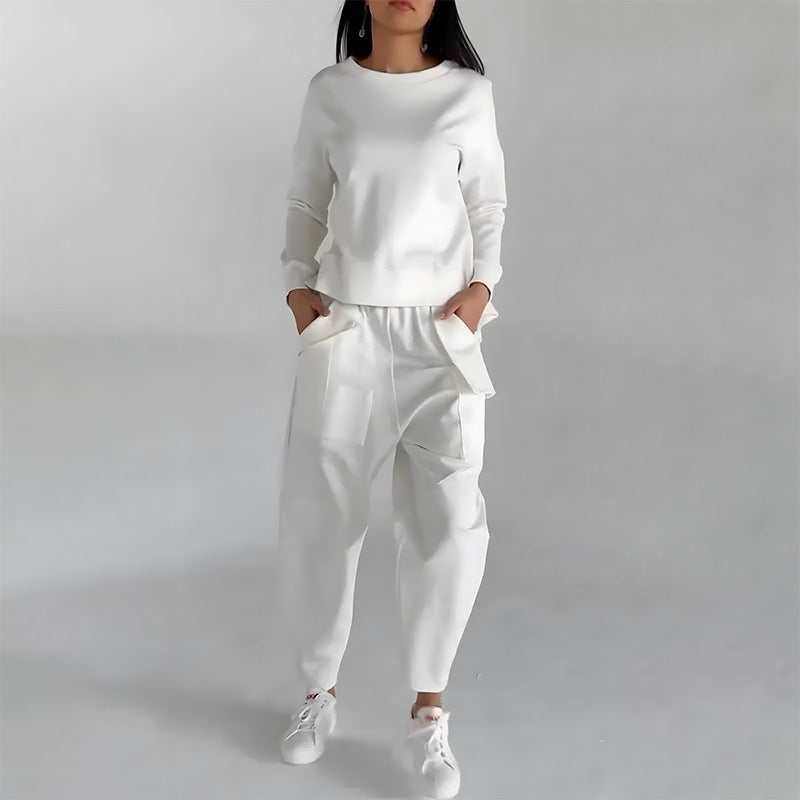 Woman wearing white fashion sweatshirt top and matching pants with hands in pockets and white sneakers against plain background