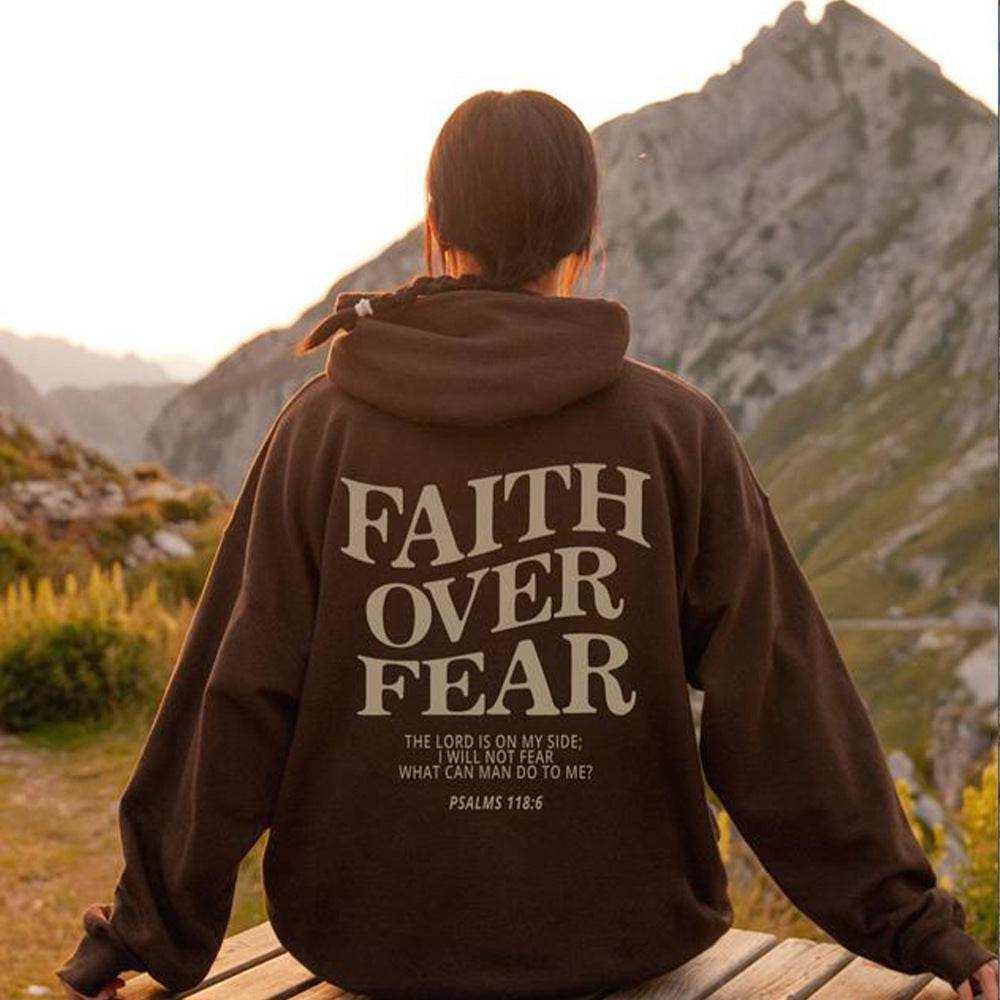 Person wearing a brown "Faith Over Fear" Christian hoodie with Psalms 118:6 text, sitting outdoors with mountain scenery in the background.